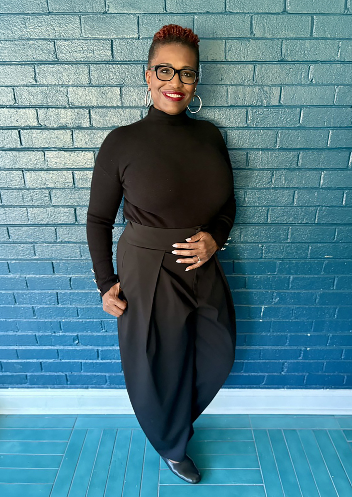 Tori O'Neal leans against a brick wall painted in a shade of deep turquoise. She is smiling and wearing a black long-sleeved turtleneck top and black pants.